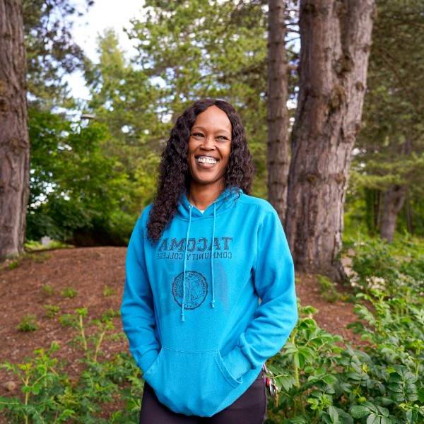 A headshot of Laquida. She smiles against a wooded background, wearing a bright blue "Tacoma Community College" sweatshirt.